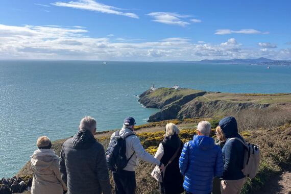 Zes mensen staan tijdens hun busrondreis Ierland op een klifpad aan de kust met uitzicht op de oceaan, jassen en rugzakken aan, starend naar een vuurtoren in de verte boven op een rotsachtige landtong omringd door weelderig groen onder een blauwe hemel en zonlicht. | Echt Ierland Vakanties