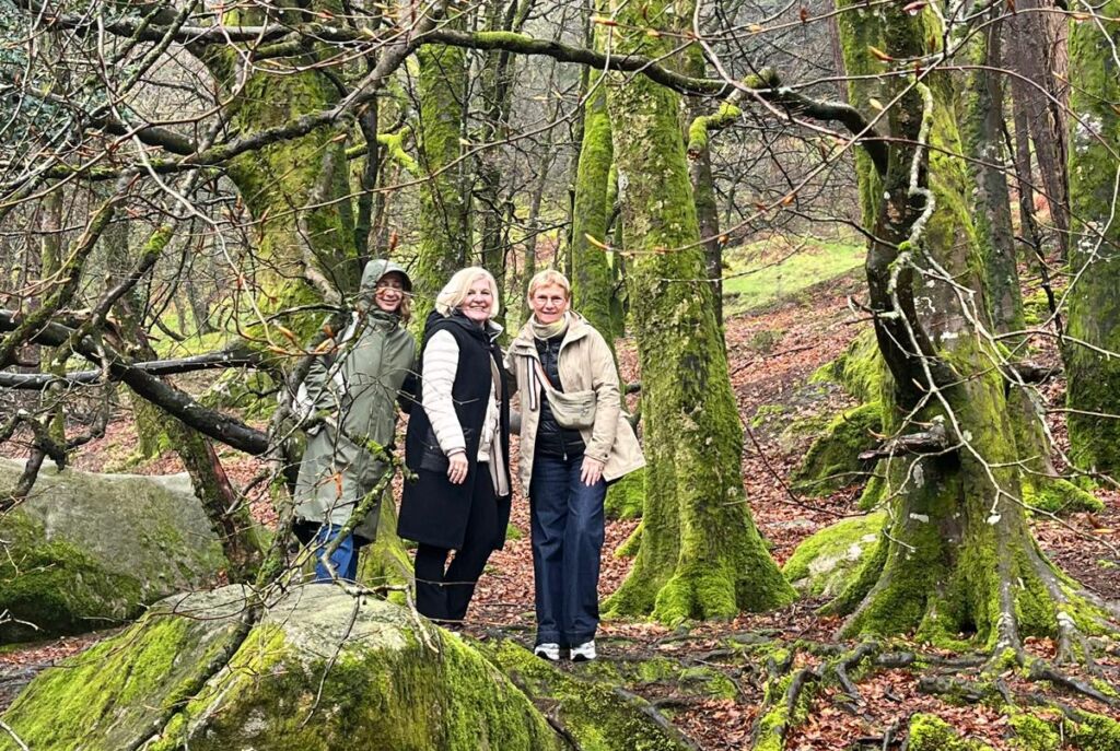 Drie mensen staan glimlachend in een weelderig, bemost Iers bos, felgroen mos dat rotsen en boomstammen bedekt. Ze zijn warm gekleed en genieten van het serene, mistige landschap - een onvergetelijke stop tijdens hun busrondreis Ierland. | Echt Ierland Vakanties