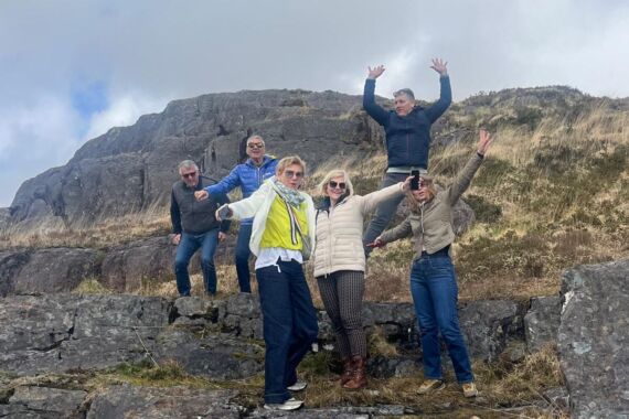 Vijf volwassenen poseren vrolijk op een rotsachtig terrein met grasvlakten tijdens een busrondreis Ierland. Twee vooraan dragen lichte jassen terwijl de anderen achteraan hun handen opsteken. Een bewolkte lucht en een rotsachtige heuvel maken deze dynamische, winderige wandelscène compleet. | Echt Ierland Vakanties