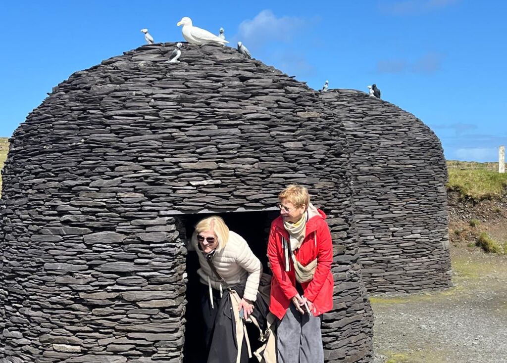 Twee vrouwen staan bij de ingang van een kleine, ronde stenen hut tijdens een busrondreis Ierland; de ene leunt glimlachend voorover terwijl de andere in een rode jas vlakbij staat. Gebeeldhouwde witte vogels toppen de hut tegen een helderblauwe lucht met verspreide wolken. | Echt Ierland Vakanties