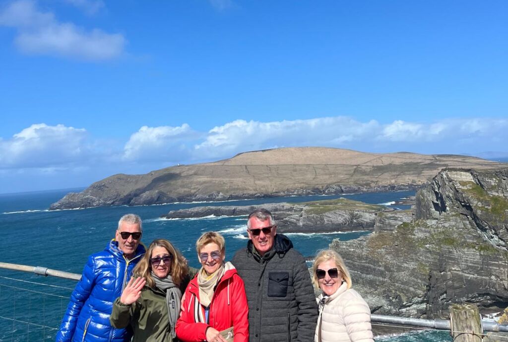 Vijf volwassenen in winterjassen staan glimlachend op een klif aan de kust onder een helderblauwe hemel. Achter hen creëren ruige kliffen en golven die tegen de rotsen slaan een prachtig tafereel - perfect voor een busrondreis Ierland avontuur. | Echt Ierland Vakanties