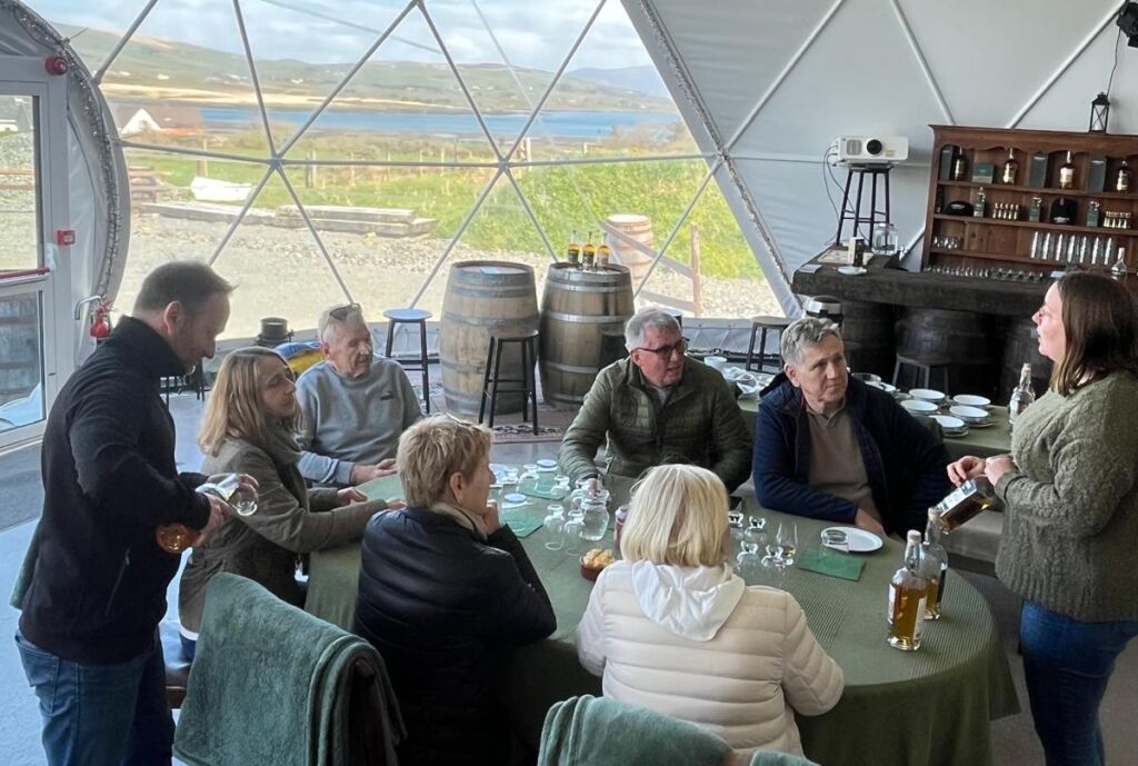 Een groep van acht mensen, waarschijnlijk op een busrondreis Ierland, verzamelt zich rond een ronde tafel in een koepelvormige kamer met grote ramen die uitkijken op schilderachtige Ierse heuvels en water. Een vrouw schenkt drankjes in terwijl anderen toekijken, omringd door rustieke vaten, planken en flessen. | Echt Ierland Vakanties