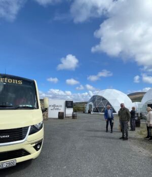 Een crèmekleurige tourbus met de naam Bartons, onderdeel van busrondreis Ierland, staat geparkeerd bij geodetische koepeltenten. Verschillende mensen staan of lopen op het grindveld onder een blauwe lucht met pluizige wolken. Vaten en bewegwijzering liggen verspreid, met heuvels op de achtergrond. | Echt Ierland Vakanties