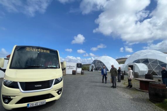 Een crèmekleurige tourbus met de naam Bartons, onderdeel van busrondreis Ierland, staat geparkeerd bij geodetische koepeltenten. Verschillende mensen staan of lopen op het grindveld onder een blauwe lucht met pluizige wolken. Vaten en bewegwijzering liggen verspreid, met heuvels op de achtergrond. | Echt Ierland Vakanties