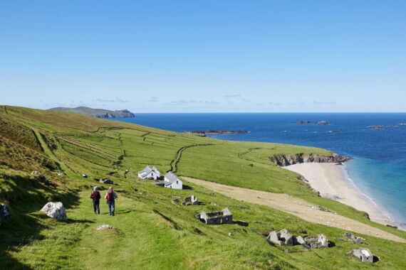 Twee mensen lopen over een met gras begroeid heuvelpad in de richting van witte huizen, met uitzicht op een zandstrand en een blauwe oceaan onder een heldere hemel - een perfect tafereel voor vakantie-ierland-2026. Stenen ruïnes stippelen het landschap uit en groene velden strekken zich uit tot verre heuvels. | Echt Ierland Vakanties