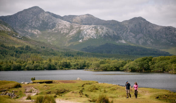 Twee mensen lopen over een graspad bij een meer, omringd door dichte groene bomen en hoge, rotsachtige bergen onder een bewolkte hemel. De scène legt de vredige schoonheid van vakantie-ierland-2026 vast, perfect voor een schilderachtige wandeling en ontspanning in de natuur. | Echt Ierland Vakanties