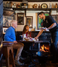Een serveerster overhandigt borden aan een man en vrouw aan een gezellige houten tafel in een rustieke pub. De kamer ademt de sfeer van vakantie-ierland-2026, met een gloeiende open haard, vintage decor, ingelijste foto's en planken vol flessen en aardewerk. | Echt Ierland Vakanties