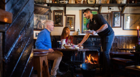 Een serveerster overhandigt borden aan een man en vrouw aan een gezellige houten tafel in een rustieke pub. De kamer ademt de sfeer van vakantie-ierland-2026, met een gloeiende open haard, vintage decor, ingelijste foto's en planken vol flessen en aardewerk. | Echt Ierland Vakanties