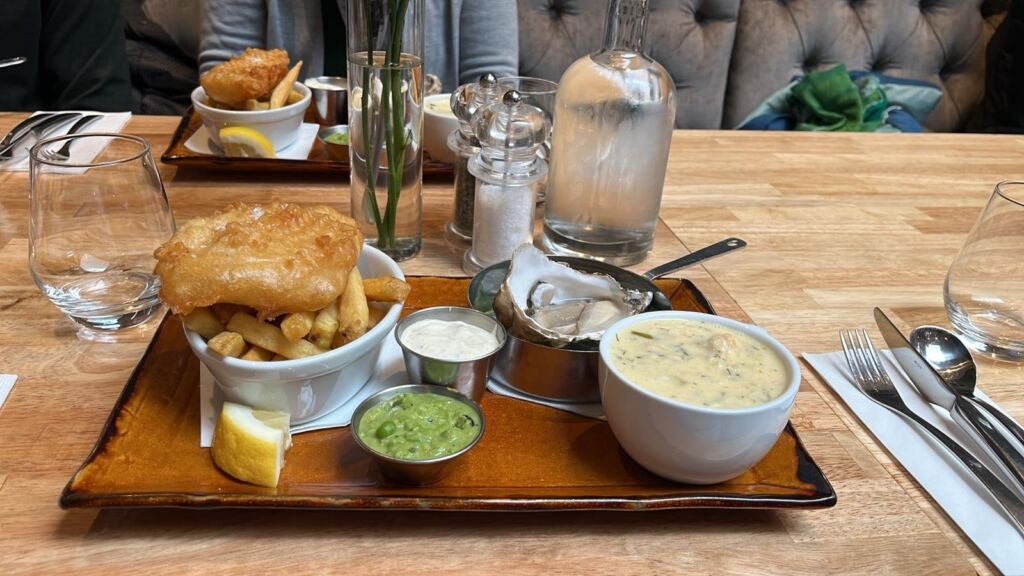Een houten gedekte tafel met een dienblad met fish and chips, mushy peas, tartaarsaus, een citroenpartje, oesters op ijs, clam chowder in een kom en een glazen waterfles. Ongebruikt bestek, glazen en peper- en zoutstel zijn ook zichtbaar. | Echt Ierland Vakanties