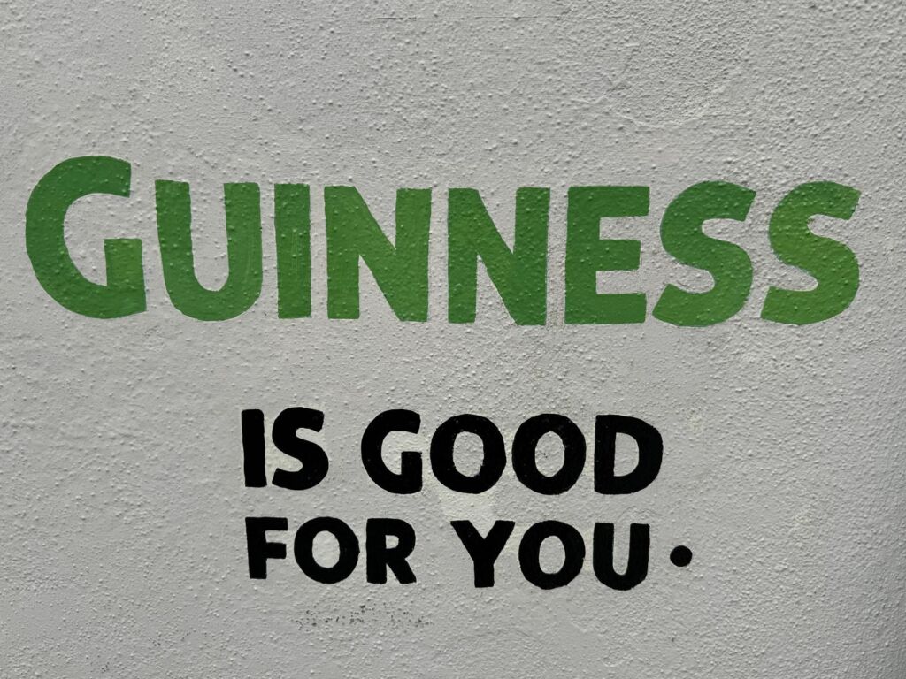 Tekst geschilderd op een ruwe, lichtgrijze muur luidt: GUINNESS in grote groene letters, boven IS GOED VOOR JE. in kleinere vette zwarte letters, met een zwarte punt aan het eind. | Echt Ierland Vakanties