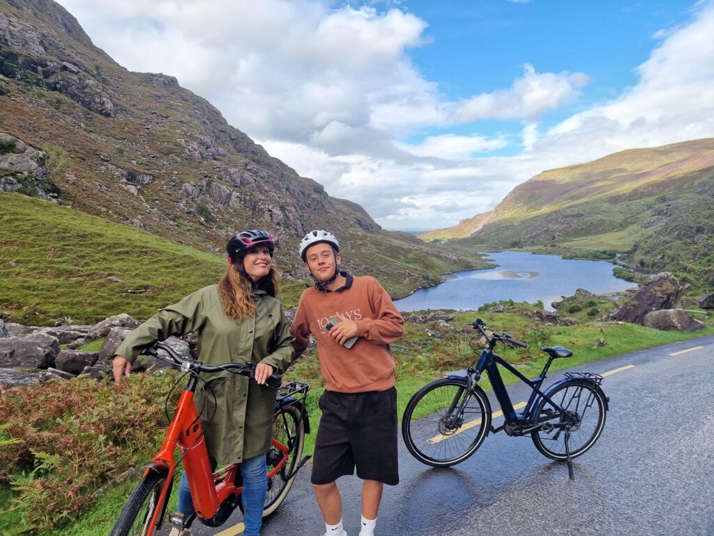 Twee mensen met fietsen poseren op een schilderachtige bergweg langs een kronkelende rivier. De vrouw draagt een groene jas en helm, glimlacht en houdt haar oranje fiets vast; de man met helm, bruine sweater en korte broek glimlacht naast een zwarte fiets. Groene heuvels en blauwe lucht vullen de achtergrond. | Echt Ierland Vakanties