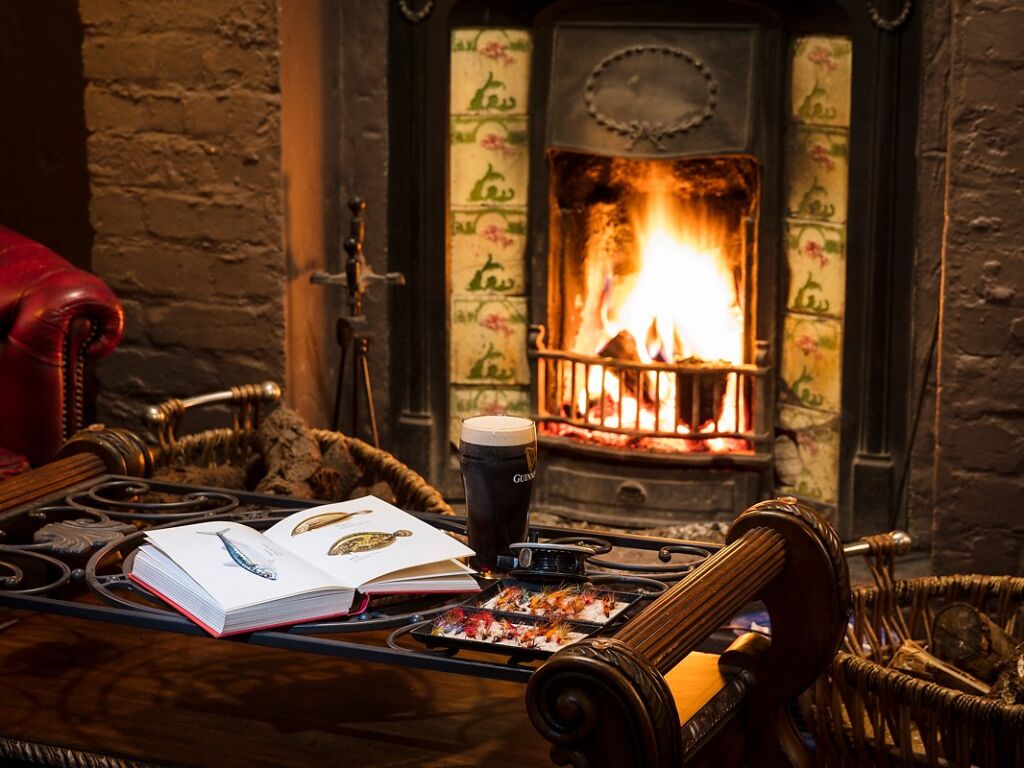 Een gezellige kamer met een brandende open haard, een sierlijke haardplaat en een houten tafel. Op de tafel staan een pint Guinness, een opengeslagen boek met visillustraties en een doos bonbons. Een roodleren stoel en rieten manden maken het warme, uitnodigende tafereel compleet. | Echt Ierland Vakanties