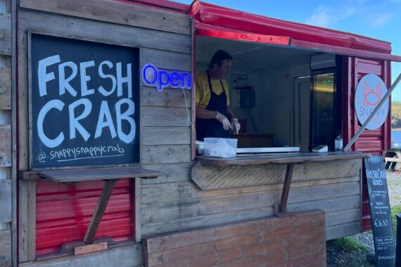 Een houten foodtruck met een bordje FRESH CRAB en een klein neon open bordje. Een persoon in een geel shirt staat binnen. Een menu op krijtbord staat bij het raam. De lucht is blauw met verspreide wolken en de grond is bedekt met grind. | Echt Ierland Vakanties