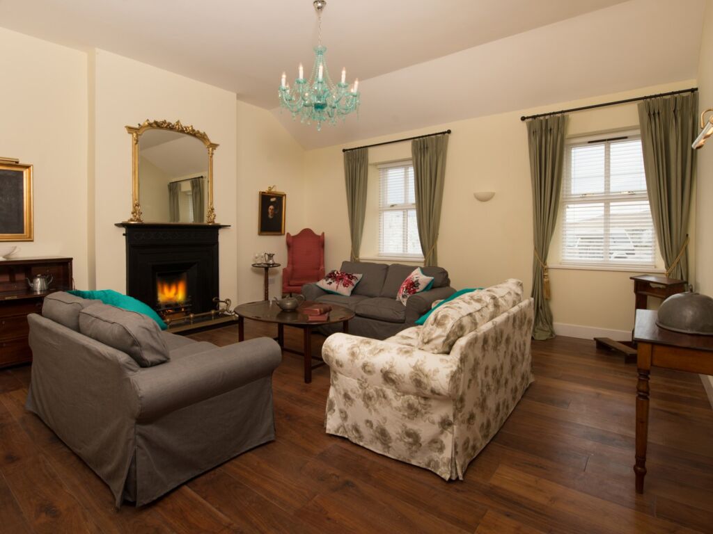 Een gezellige woonkamer met twee grijze banken en een bank met bloemmotief rond een houten salontafel, Levendig groene gordijnen voor twee ramen, een verlichte open haard met een goudomrande spiegel erboven, houten vloeren en een blauwe kroonluchter boven je hoofd. | Echt Ierland Vakanties