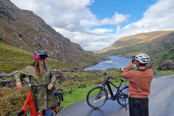 Twee fietsers met een helm op staan even stil op een bergweg; één van hen fotografeert een schilderachtig uitzicht met een rivier die zich onder een blauwe lucht met wolken een weg baant door groene heuvels. Hun fietsen staan naast hen en het weelderige landschap strekt zich uit tot in de verte. | Echt Ierland Vakanties