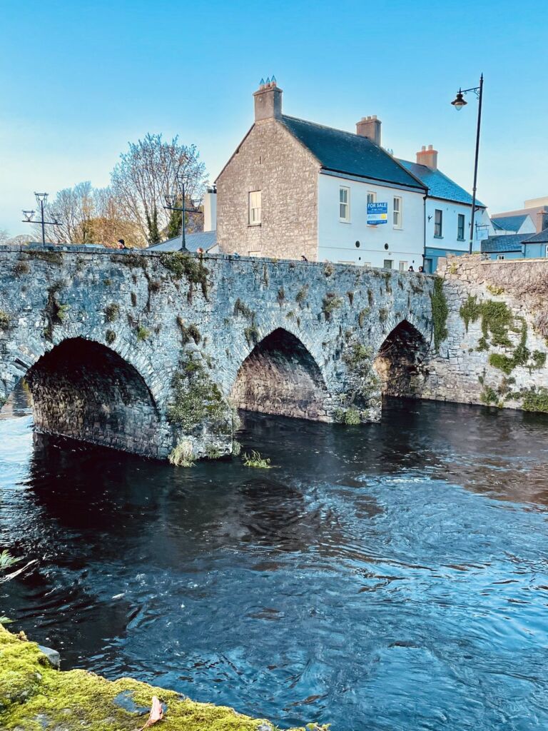Een stenen brug met drie bogen overspant een donkere, rustig stromende rivier. Aan de overkant staat vlak bij de brug een stenen huis van twee verdiepingen met wit omrande ramen en een blauw bord. Onder een helderblauwe hemel zijn bomen en een straatlantaarn te zien. | Echt Ierland Vakanties