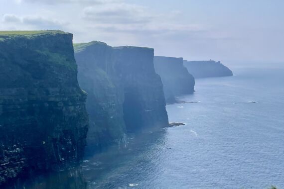 Een mistig uitzicht op de Cliffs of Moher in Ierland, met steile, ruige kliffen die boven de Atlantische Oceaan uittorenen. Zacht zonlicht laat de groene toppen van de kliffen schitteren, en het water eronder ziet er kalm uit. De kliffen strekken zich uit tot in de wazige verte onder een gedeeltelijk bewolkte hemel. | Echt Ierland Vakanties