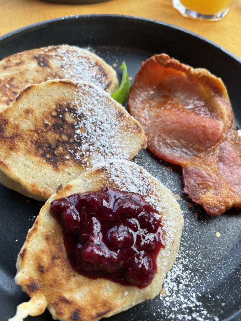 Drie dikke pannenkoeken op een zwart bord, waarvan er twee bestrooid zijn met poedersuiker en één belegd is met grofgesneden rode bessenjam. Naast de pannenkoeken liggen twee plakjes knapperig, goudbruin gebakken spek. Op de achtergrond is gedeeltelijk een glas sinaasappelsap te zien. | Echt Ierland Vakanties