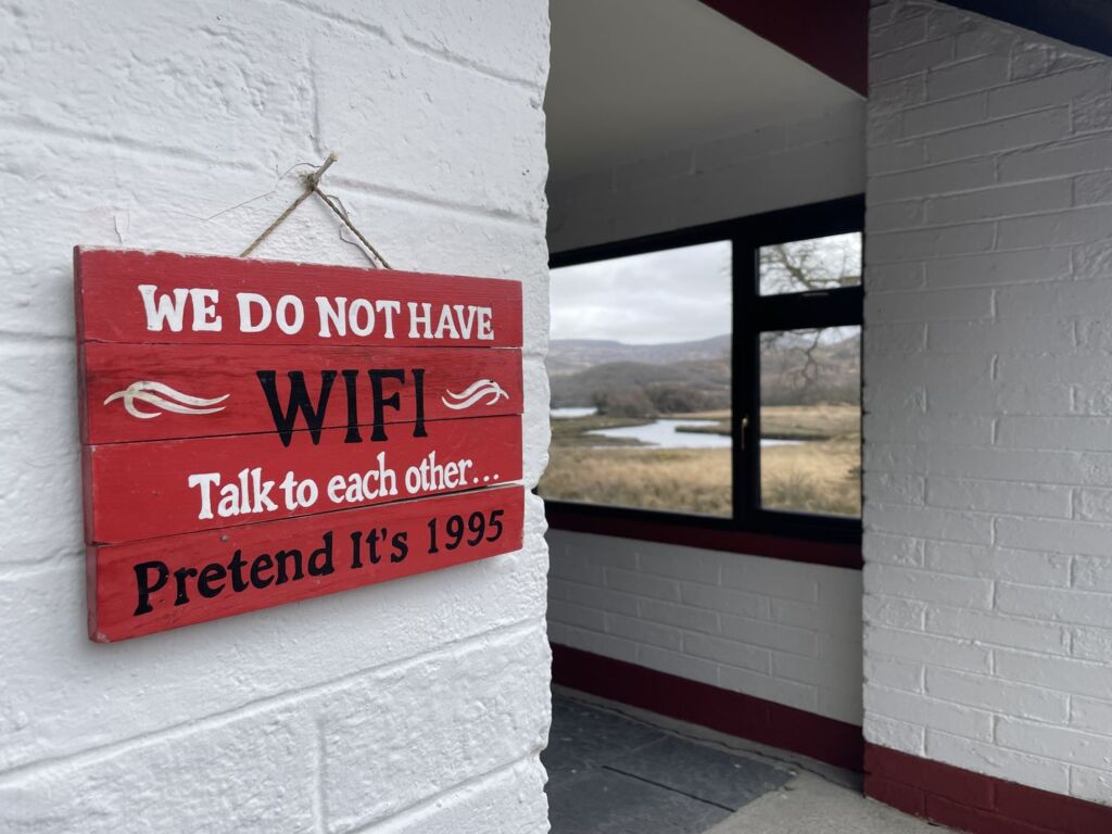 Op een wit bakstenen muur hangt een rood-wit bordje met de tekst: „WIJ HEBBEN GEEN WIFI! Praat gewoon met elkaar... Doe alsof het 1995 is.” Op de achtergrond is door een raam een schilderachtig landschap te zien met water en heuvels. | Echt Ierland Vakanties