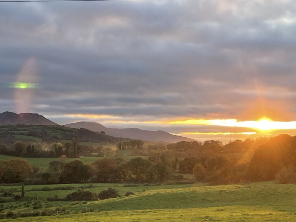 Een schilderachtig landschap bij zonsondergang, met groene velden, hier en daar een boom, glooiende heuvels en bergen in de verte. De zon staat laag aan de horizon en werpt een warme oranje gloed en lichtstralen over de gedeeltelijk bewolkte hemel. | Echt Ierland Vakanties