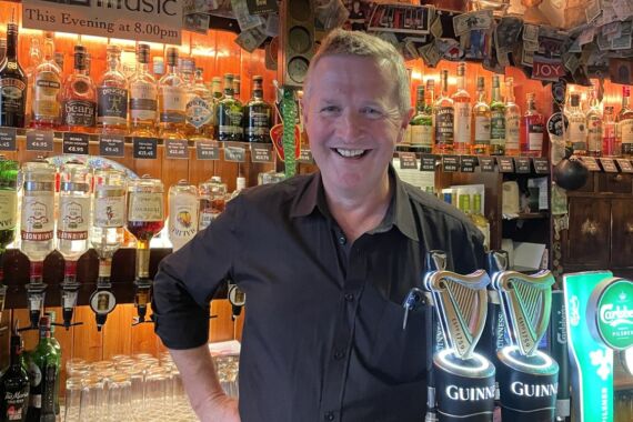 Achter de bar van een pub, met twee Guinness-taps, staat een glimlachende barman in een zwart shirt. De planken achter hem staan vol met flessen sterke drank, glazen en Ierse decoraties. Op een bord erboven staat: „Live Ierse muziek vanavond om 20.00 uur.” De bar is sfeervol verlicht. | Echt Ierland Vakanties